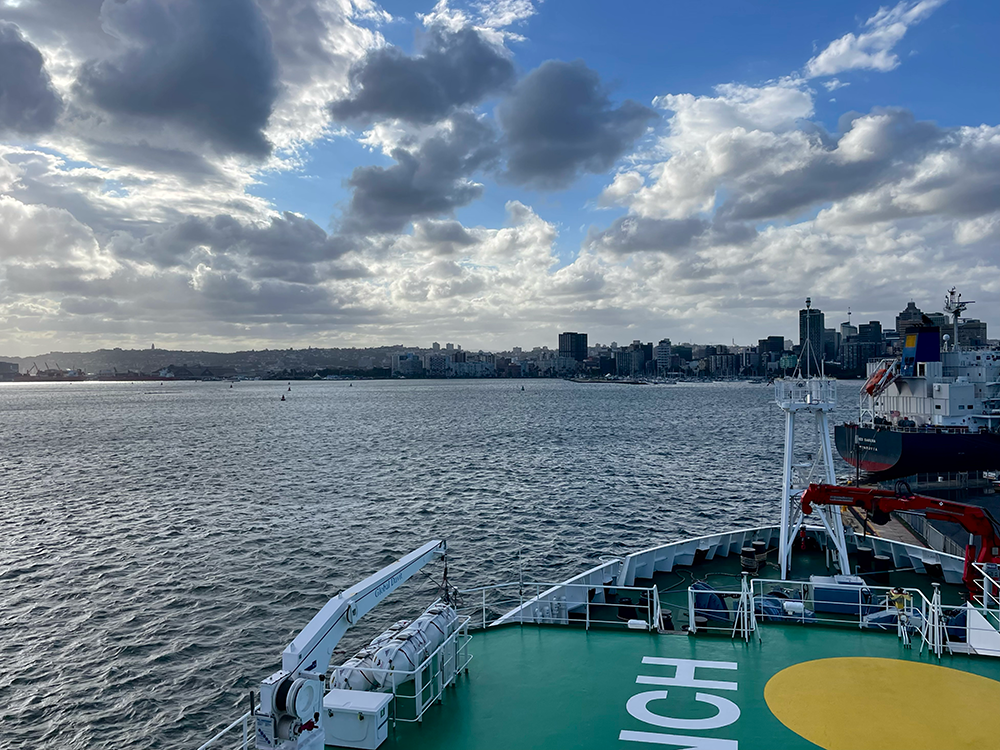 above the green bow of the ship, the skyline of Durban is visible under a cloudy sky